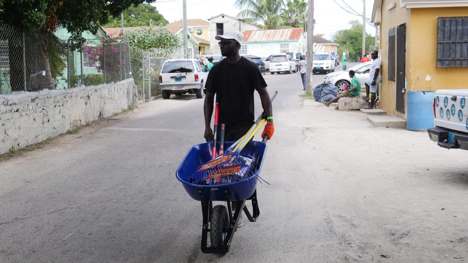 A man walking down a street pushing a blue wheelbarrow filled with cleaning supplies, wearing sunglasses and a cap.