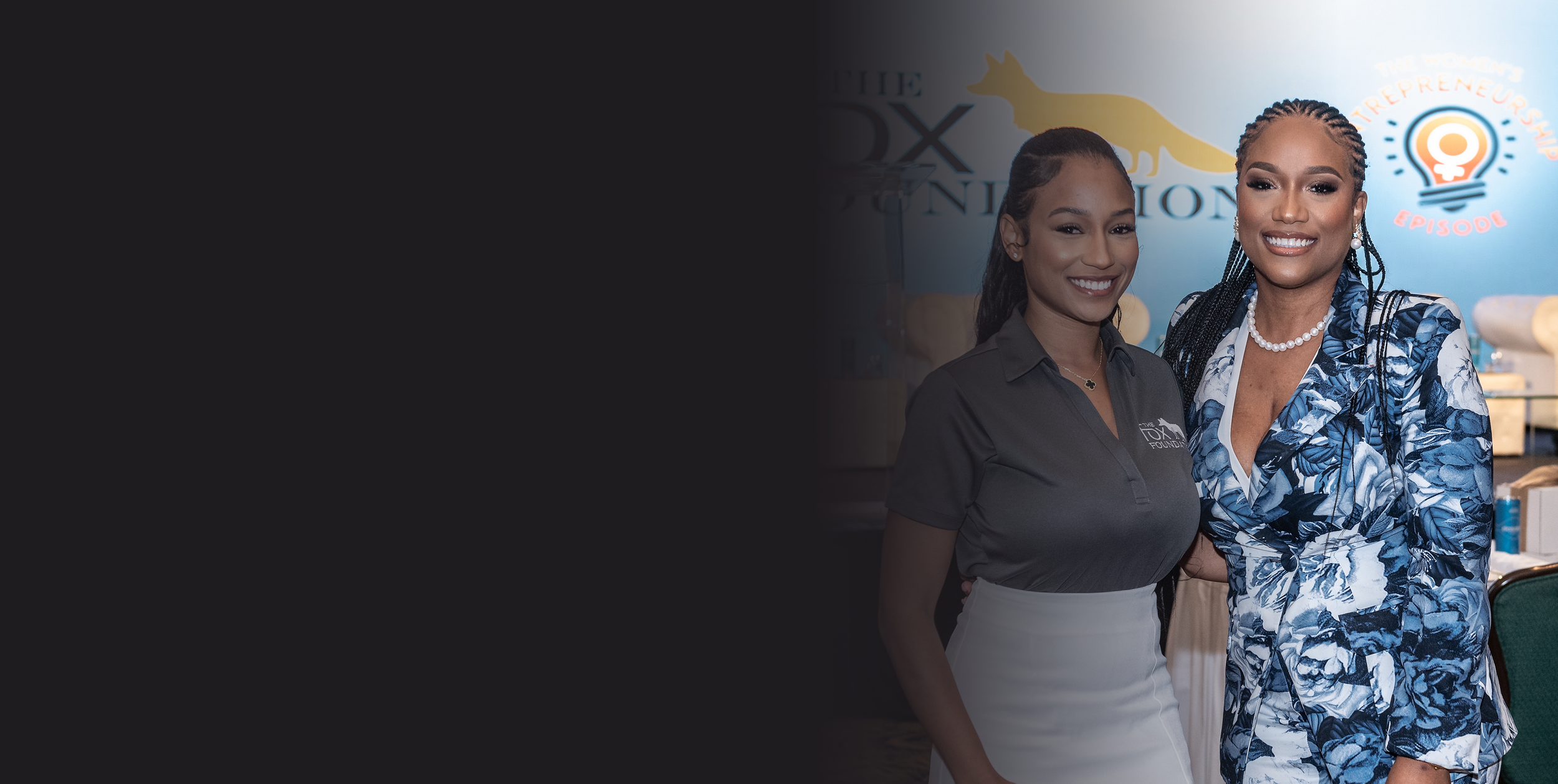 Two women smiling, standing side by side at an indoor event with a blue background displaying logos and text.