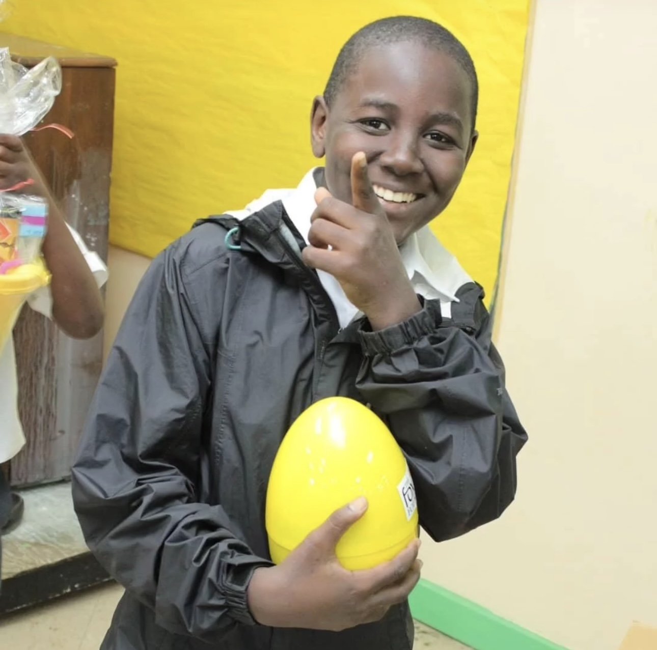 Smile of a young boy holding a yellow Easter egg, standing indoors with a yellow wall in the background.