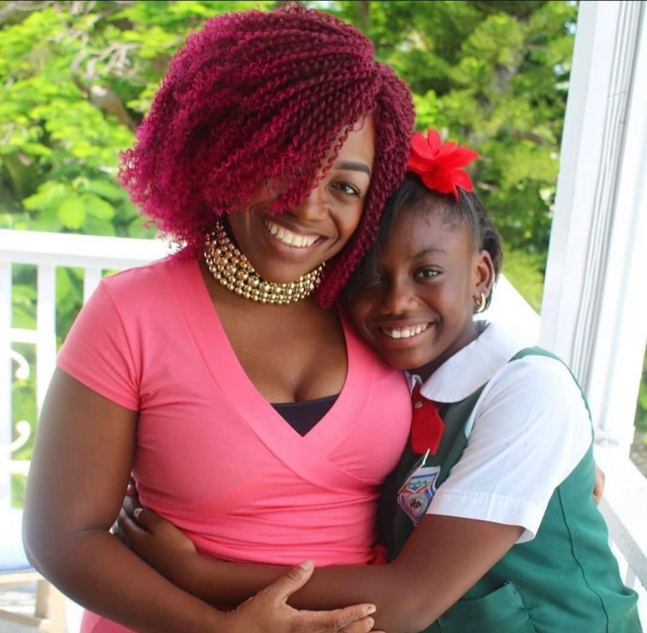 A woman and a girl are hugging and smiling outdoors. The woman has curly, red hair and is wearing a pink shirt and a gold pearl necklace. The girl has dark hair with a red bow and is wearing a white shirt with a green vest.