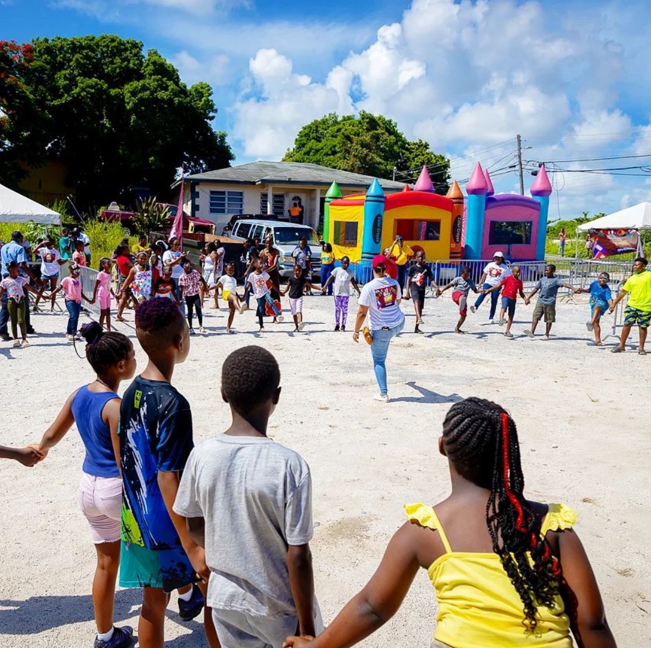 Children and adults at an outdoor community event with bounce houses, tents, and a house in the background under blue skies.