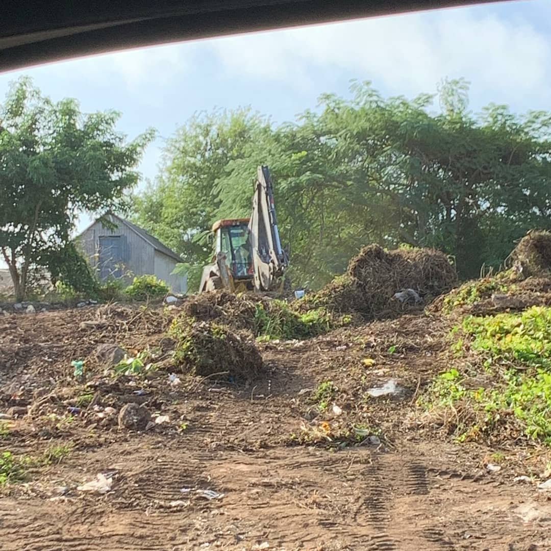 Construction site with an excavator on uneven dirt ground, surrounded by green trees and a small metal shed in the background.