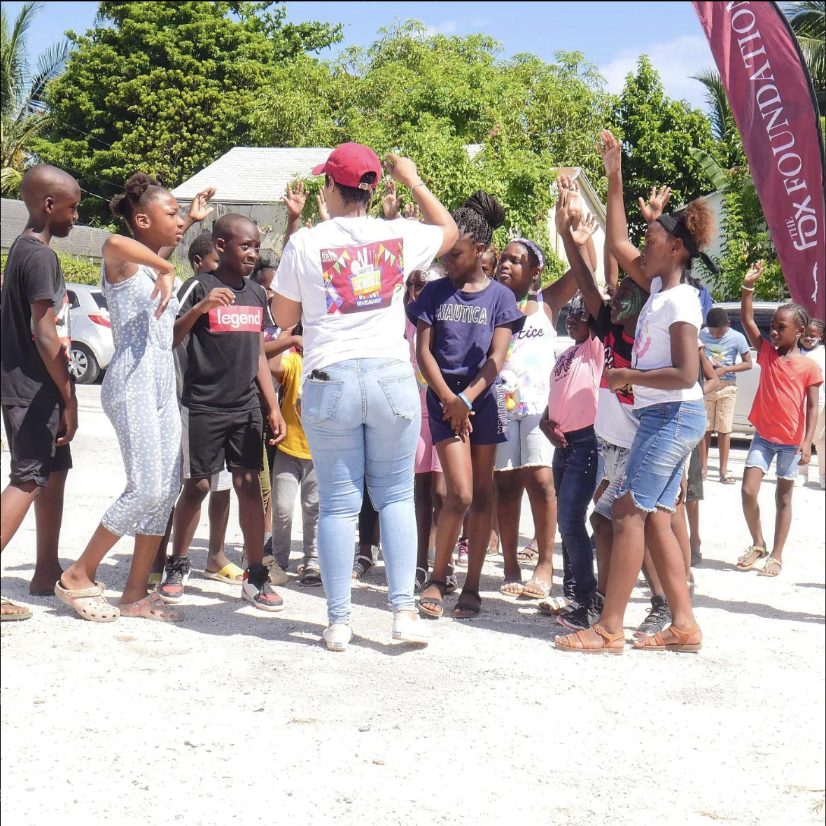 A group of children and teenagers outdoors participating in a game or activity, with some raising their hands, standing on a gravel surface, surrounded by trees and parked cars.
