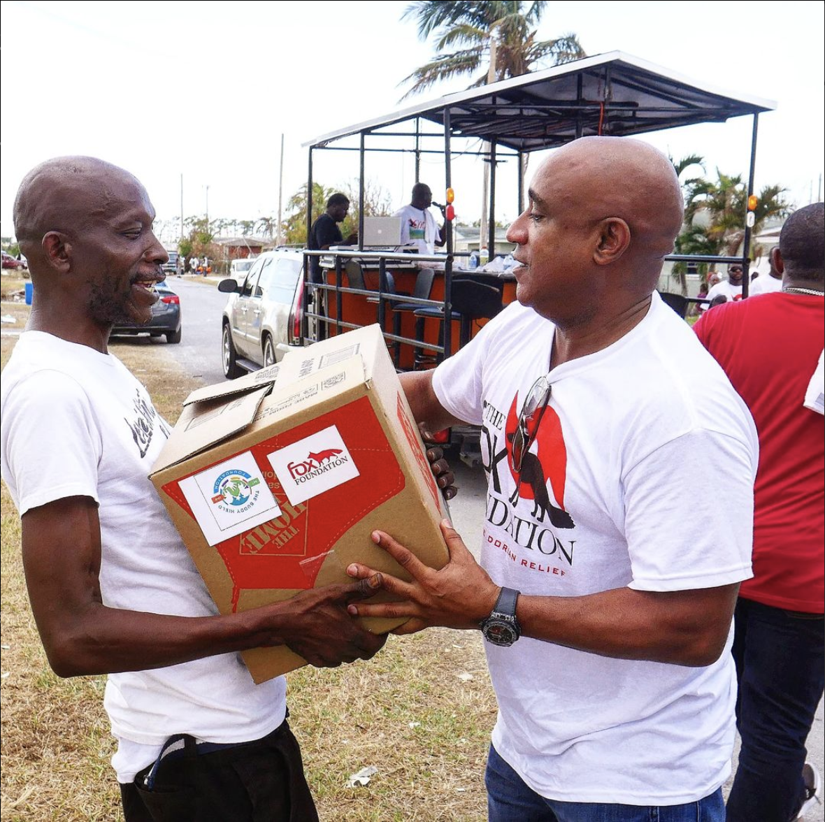 Two men smiling as one hands a cardboard box with the logos of the Fox Foundation and Food for the Hungry to the other during a charity event outdoors.