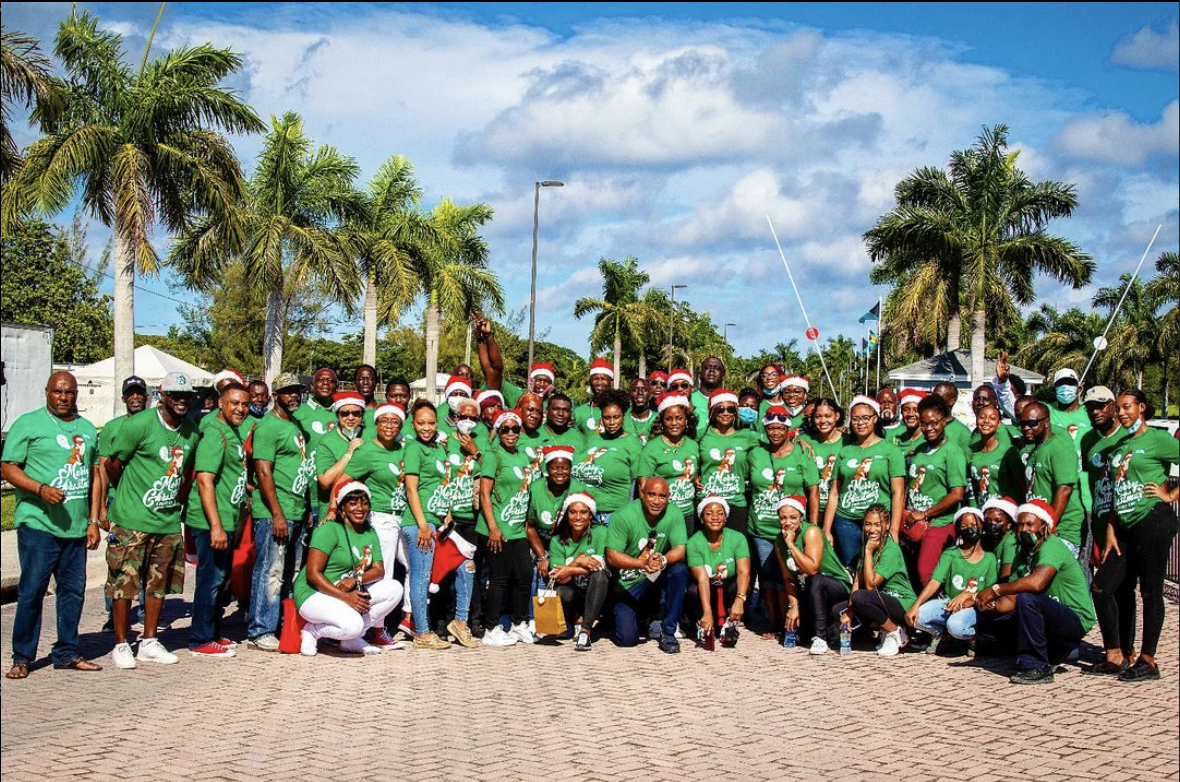Group of people wearing matching green Christmas-themed shirts and Santa hats outdoors, with palm trees and a blue sky in the background.