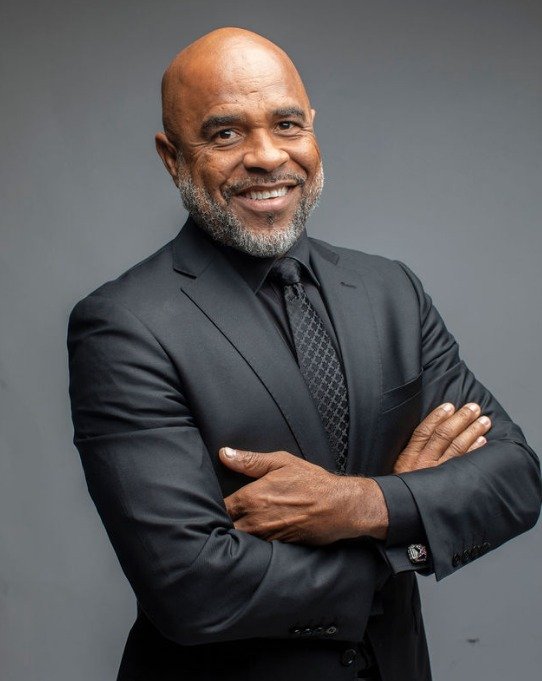A smiling bald man with a gray beard, wearing a dark suit and tie, standing with crossed arms against a gray background.
