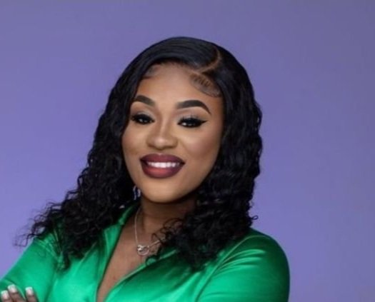 Portrait of a woman with dark, wavy hair wearing a green satin blazer, smiling against a purple background.