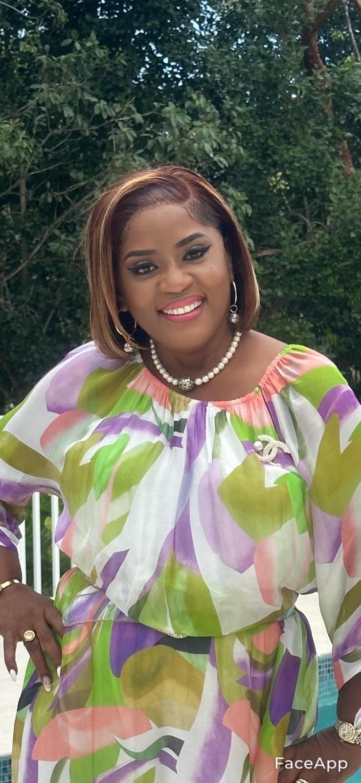 A woman with shoulder-length light brown hair, wearing makeup, jewelry including earrings, a necklace, and rings, dressed in a colorful, patterned dress with green, purple, pink, and white hues, standing outdoors in front of green foliage.