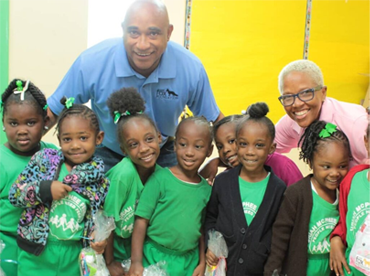 A group of diverse children in green shirts posing with The Honoroable Dr. Adrian Fox and Ms. Phillipa Ambrister in an indoor setting with yellow and blue backgrounds.