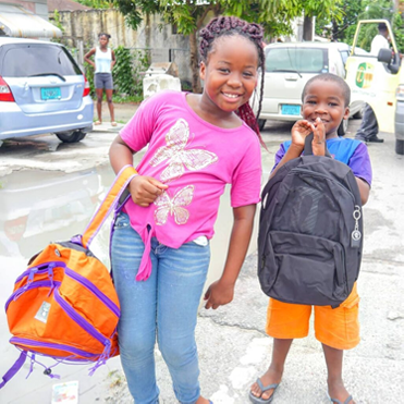Two smiling children with backpacks standing in a parking lot with cars and trees in the background.